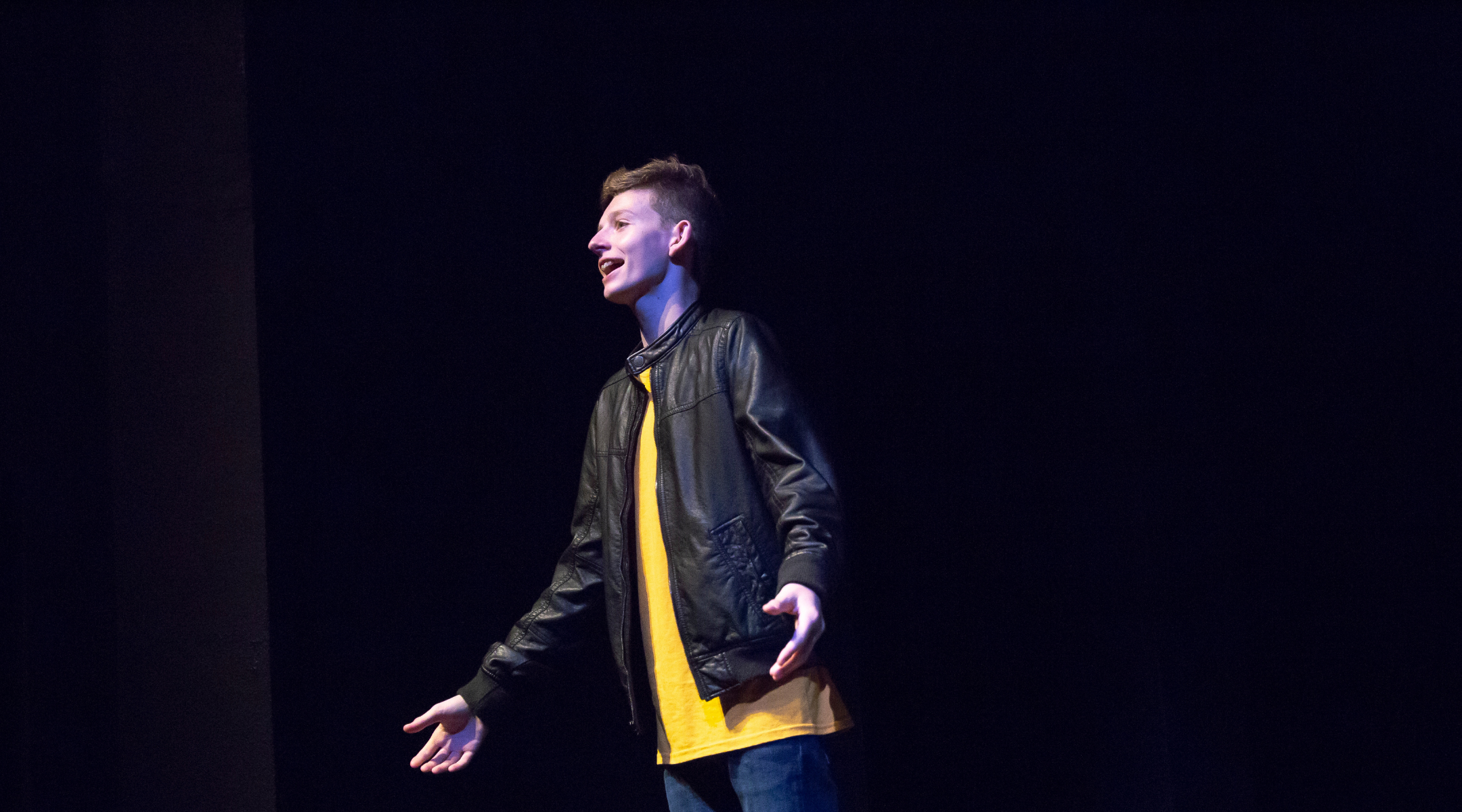 student wearing black leather jacked and yellow shirt singing on stage